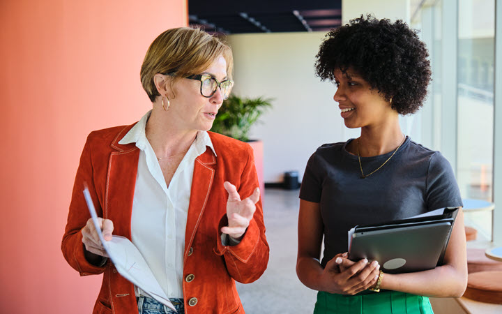 Two women standing in an office talking. One woman is holding papers; the other woman is holding a notebook and computer.