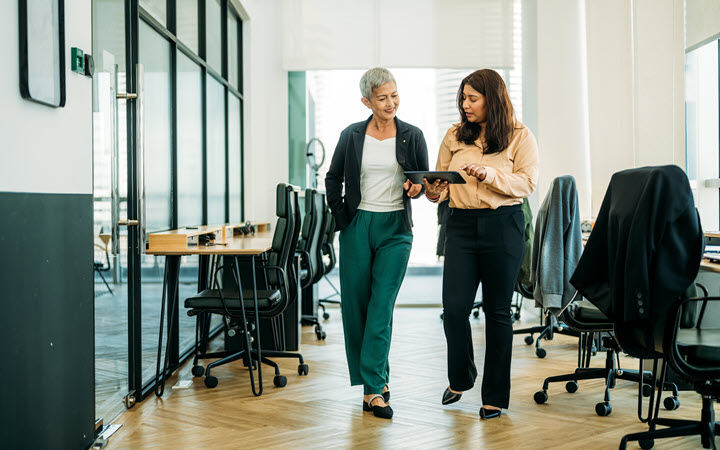 Two women walking through an office and looking at a tablet computer.