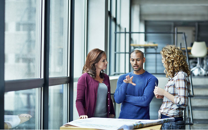 A man and two woman talking together in a office and looking at paper plans.